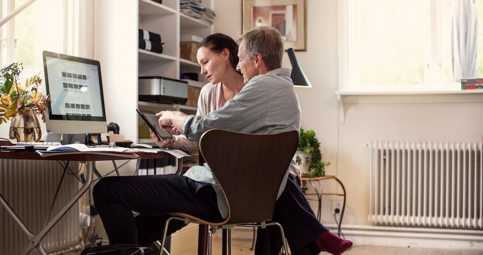 Mann und Frau sitzen in einem modernen, hellen Arbeitszimmer. Die Frau hält ein Tablet in der Hand und der Mann erklärt ihr etwas darauf. Sie hört gespannt zu.